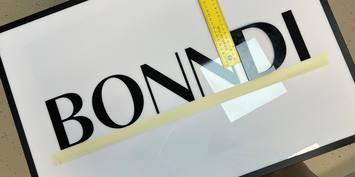 3D logo značky BONNDI s čiernymi písmenami na bielom podklade, s priloženým metrom na meranie výšky písmen.